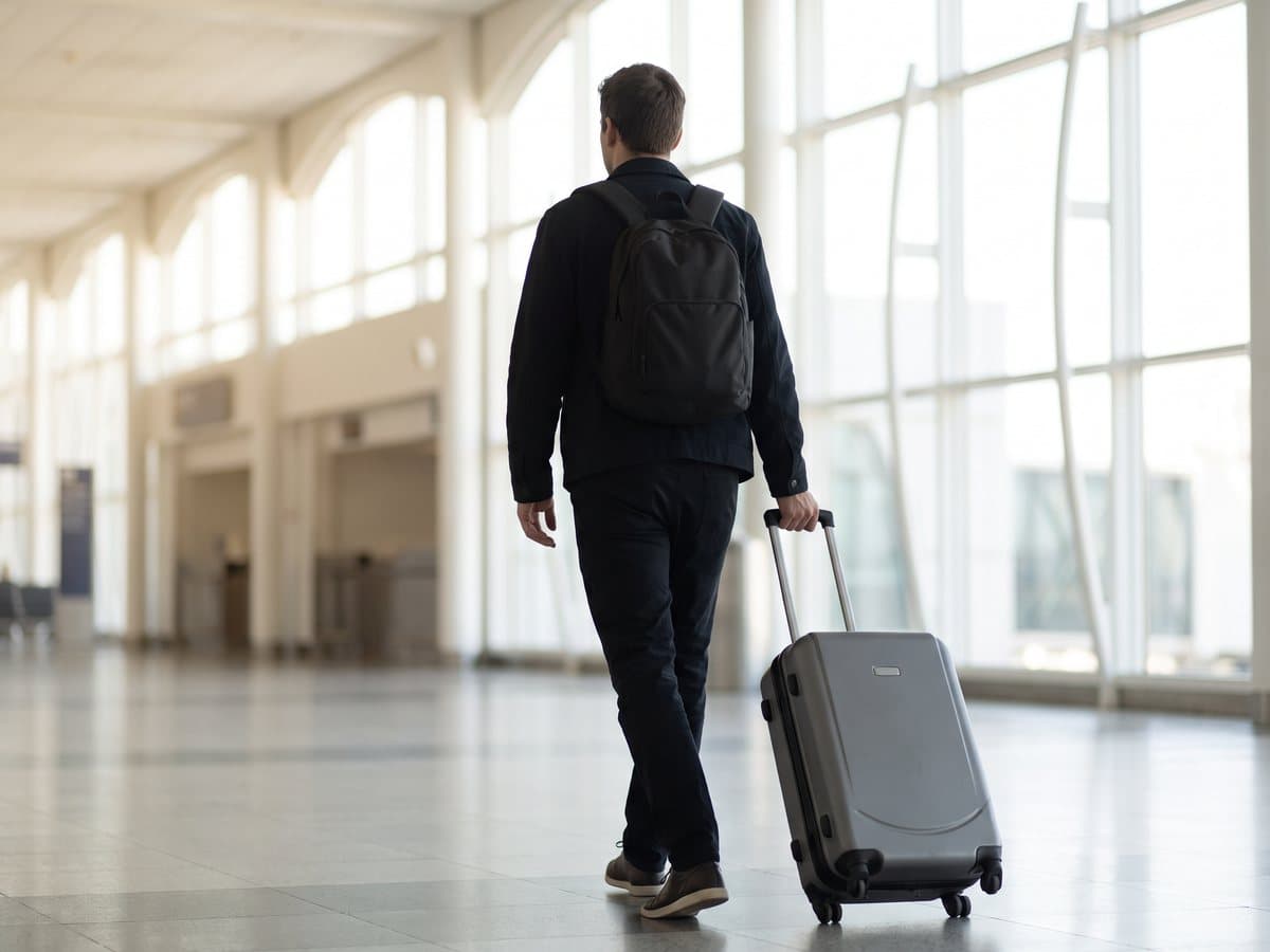 A traveler walking through a bright airport concourse pulling a hardshell carry-on spinner, a travel backpack on their shoulders