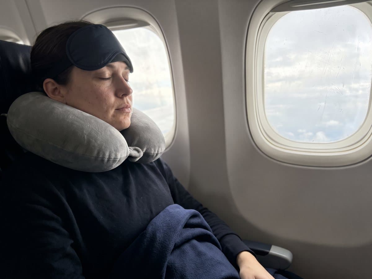 A passenger resting against an airplane window seat wearing a memory foam neck pillow and a sleep mask pushed up on the forehead, a folded travel blanket on the lap